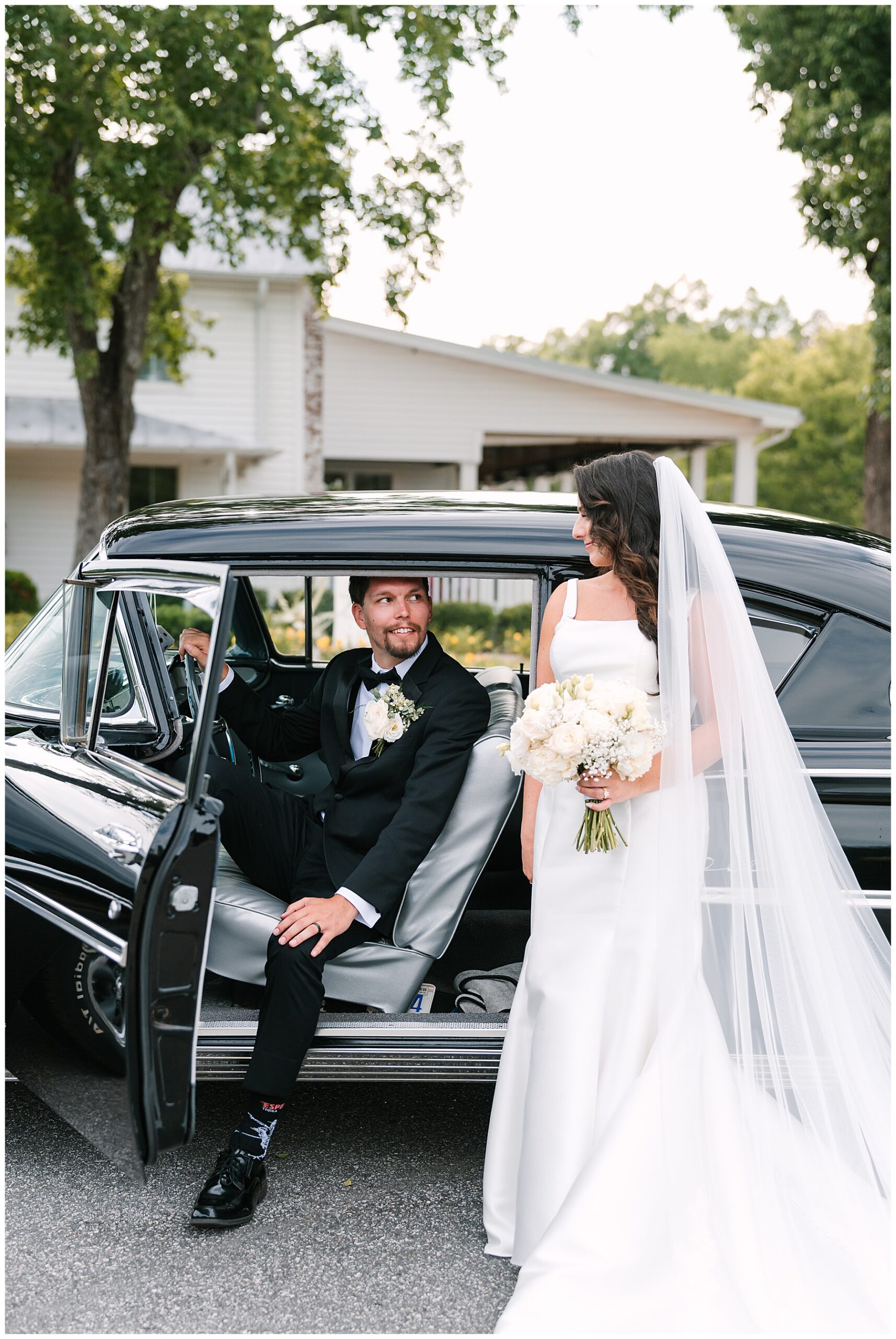 Groom sits in a classic car while his bride stands beside him outside the car door